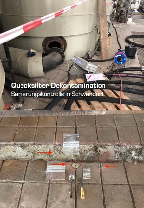 Das Bild zeigt eine Fabrikhalle und in Großaufnahme einen verkleideten Behälter mit einem abgeschraubten Rohr, das vor dem Ausgang des Behälters liegt.Das Quecksilbermessgerät, ein ca. 50cm breiter Kasten mit Digitalanzeige saugt Luft aus dem Boden vor dem Behälter. In den dicken alten Tonfliesen sieht man Bohrlöcher, das Bohrmehl wird als Probe genommen.